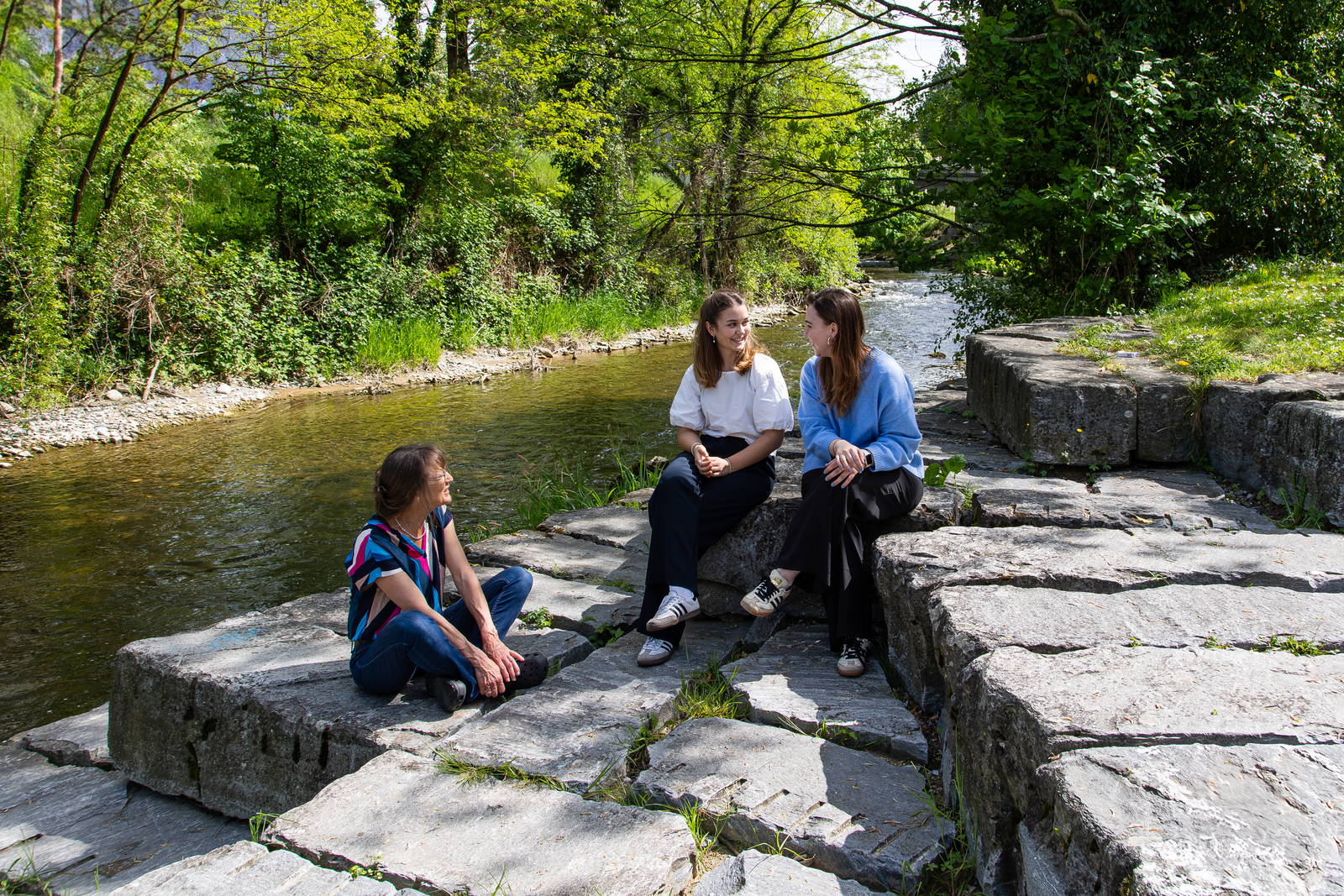 Drei Frauen sitzen im Gespräch auf Steinplatten an einem Fluss